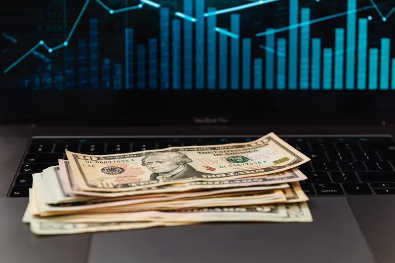 US dollar bills stacked on a laptop keyboard with a blue growth chart displayed on the screen behind, representing income potential from a faceless account