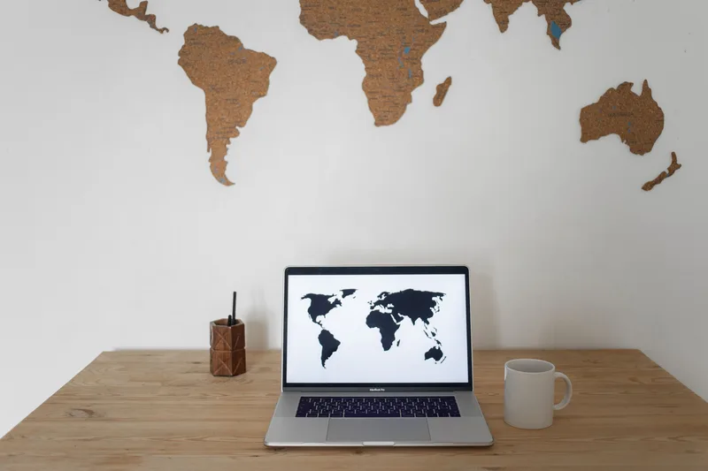A world map displayed on a laptop screen next to a ceramic cup on a desk, representing screen-based, faceless travel planning content