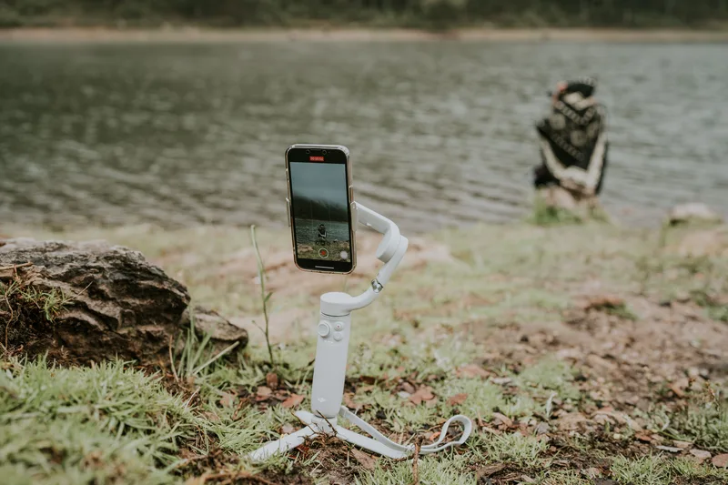 Smartphone held vertically on a white gimbal, recording a lakeside scene outdoors