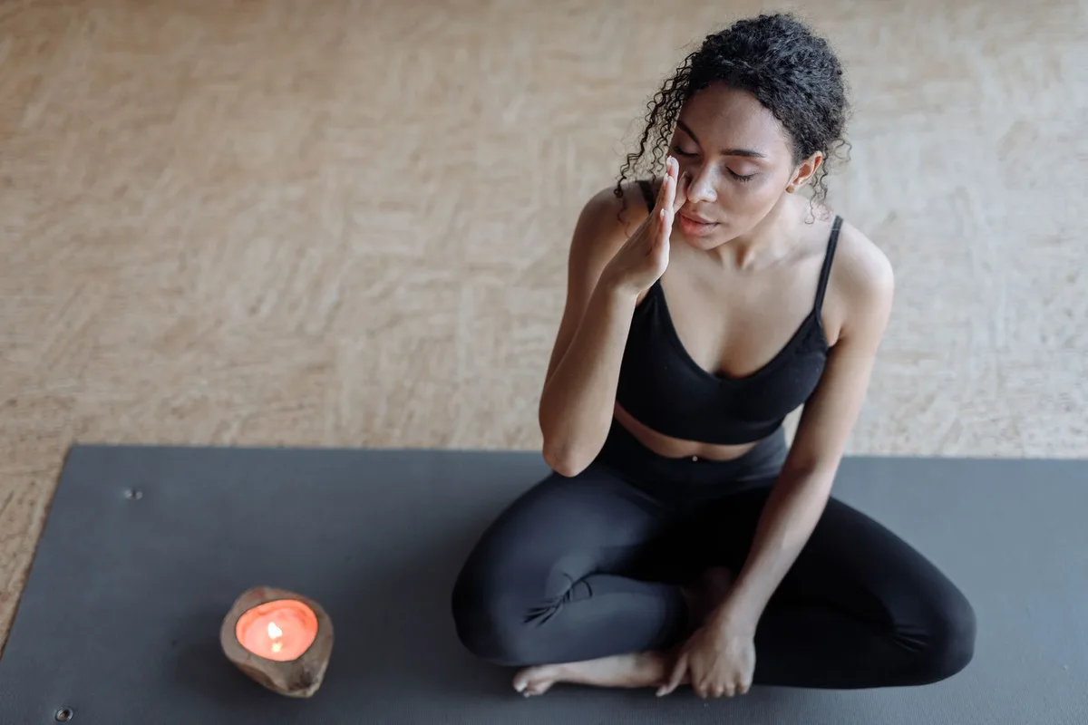 A woman practicing meditation on a yoga mat, focusing with eyes closed next to a candle indoors