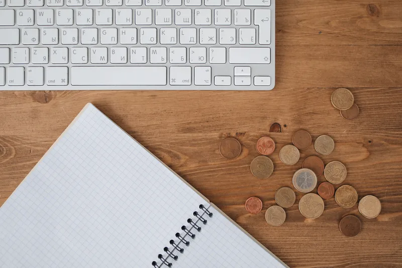 Blank grid notebook beside a white keyboard and scattered coins on a wooden desk