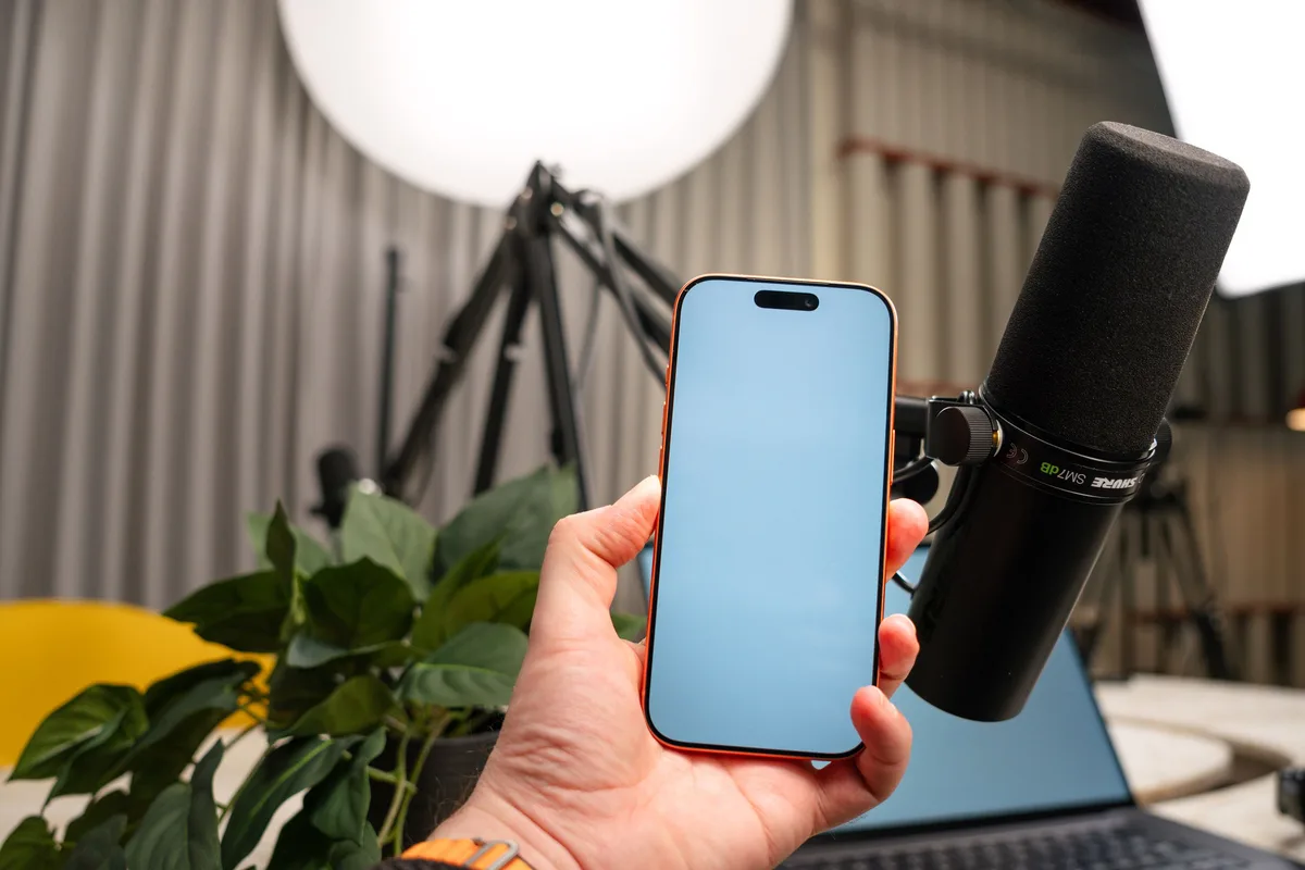 Hand holding a smartphone next to a black podcast microphone on a boom arm, with a plant and studio light behind