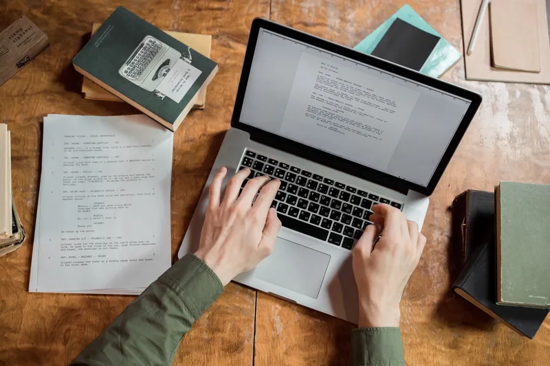 Overhead view of two hands typing on a laptop on a wooden desk surrounded by printed pages and books