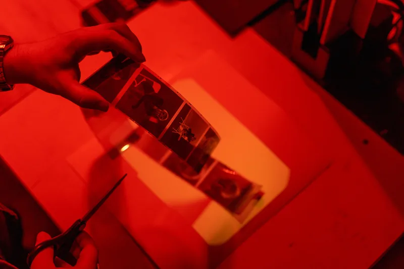 Hands holding a strip of film negatives with scissors nearby under red darkroom light, no faces visible