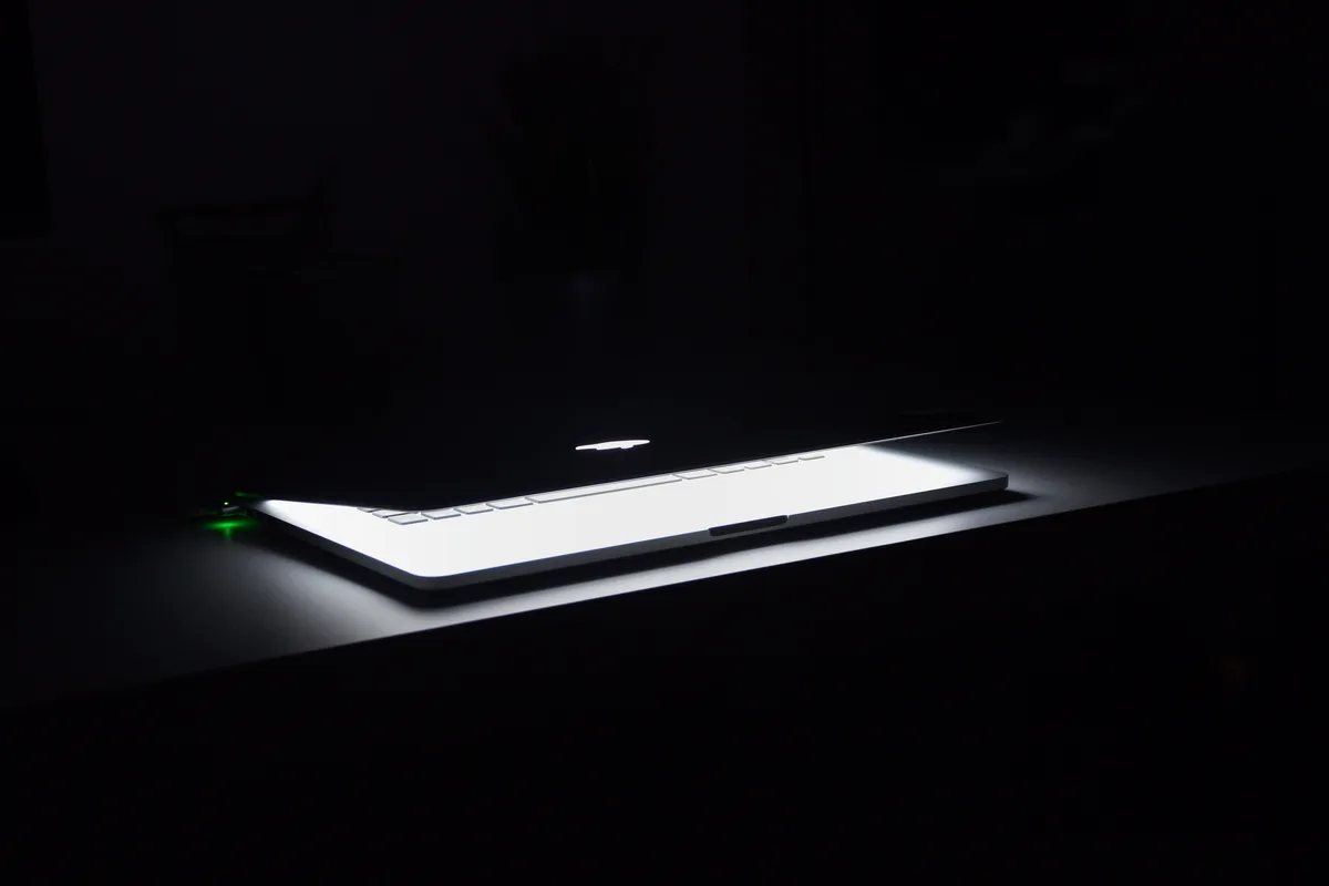 A laptop with glowing keys sitting half open on a dark desk at night, no person visible