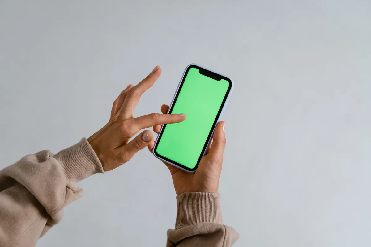 Two hands holding a smartphone upright with a blank green screen against a light gray wall, no face visible