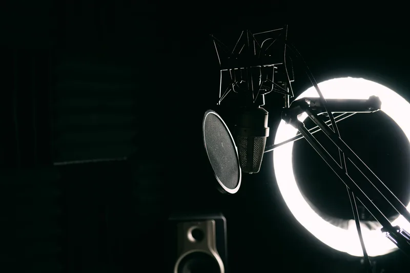 Studio microphone with a pop filter in front of a glowing ring light in a dark room, no person visible