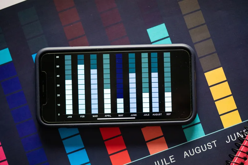 Smartphone lying sideways on a colorful printed chart, its screen showing a monthly bar chart, no person visible